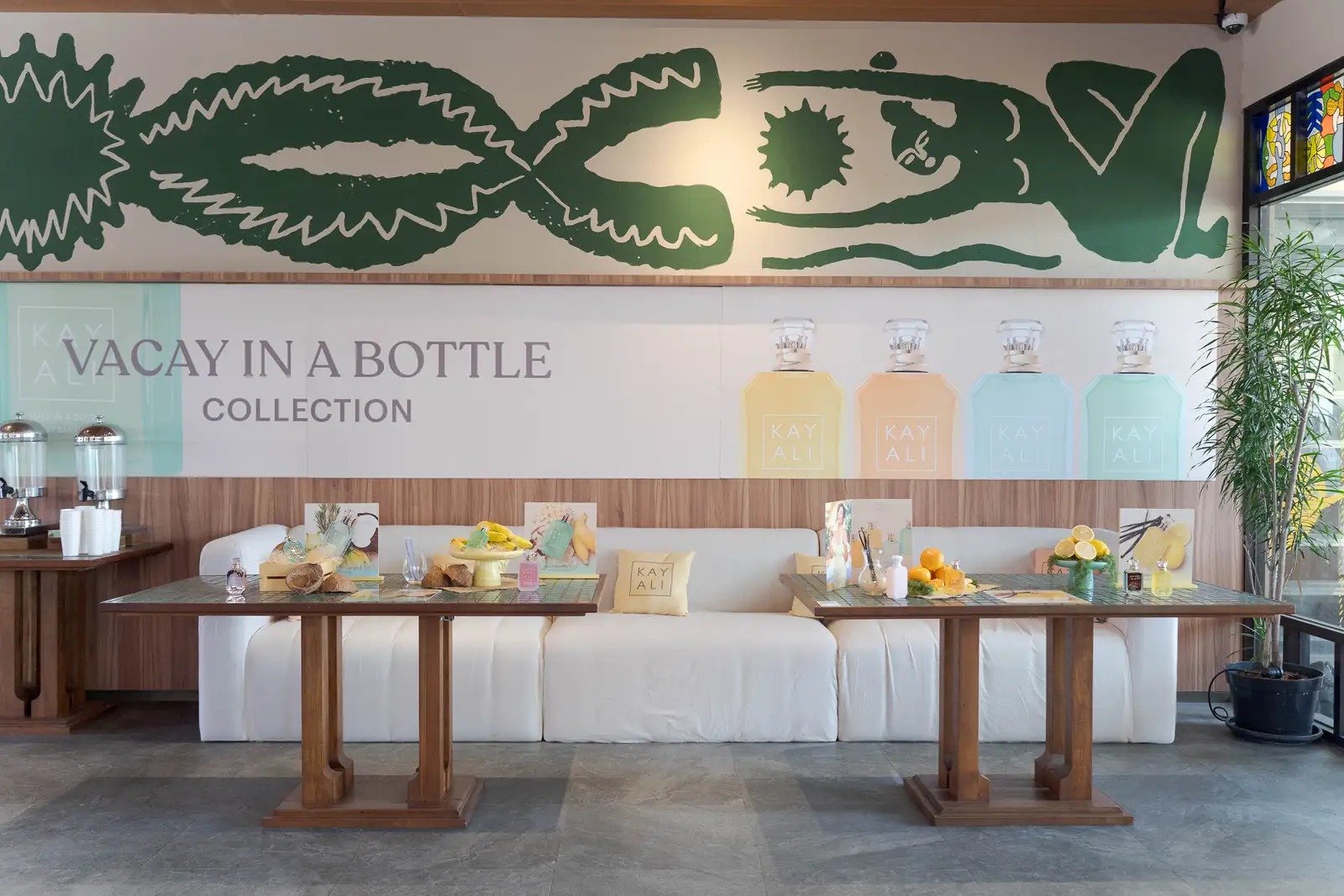 Area lounge KAYALI Vacay in a Bottle dengan mural, sofa putih, dan meja display produk di PIK 2